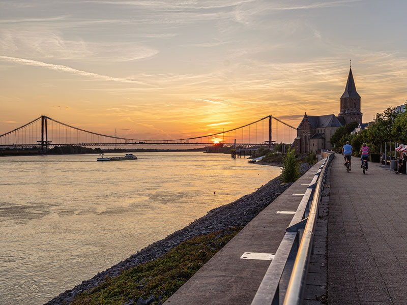 Blick auf die Rheinpromenade in Emmerich bei Sonnenuntergang mit der Rheinbrücke im Hintergrund, einer Kirche am Ufer und zwei Radfahrern auf dem Weg entlang des Flusses.