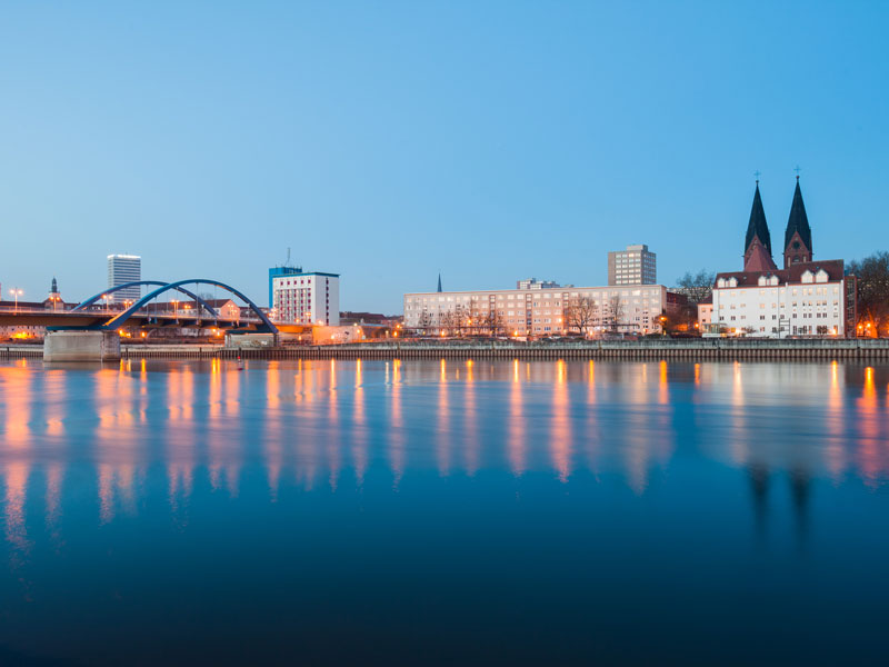 Abendliche Skyline von Frankfurt (Oder) mit Blick über die Oder auf beleuchtete Gebäude, die Stadtbrücke und die Marienkirche mit ihren Doppeltürmen.