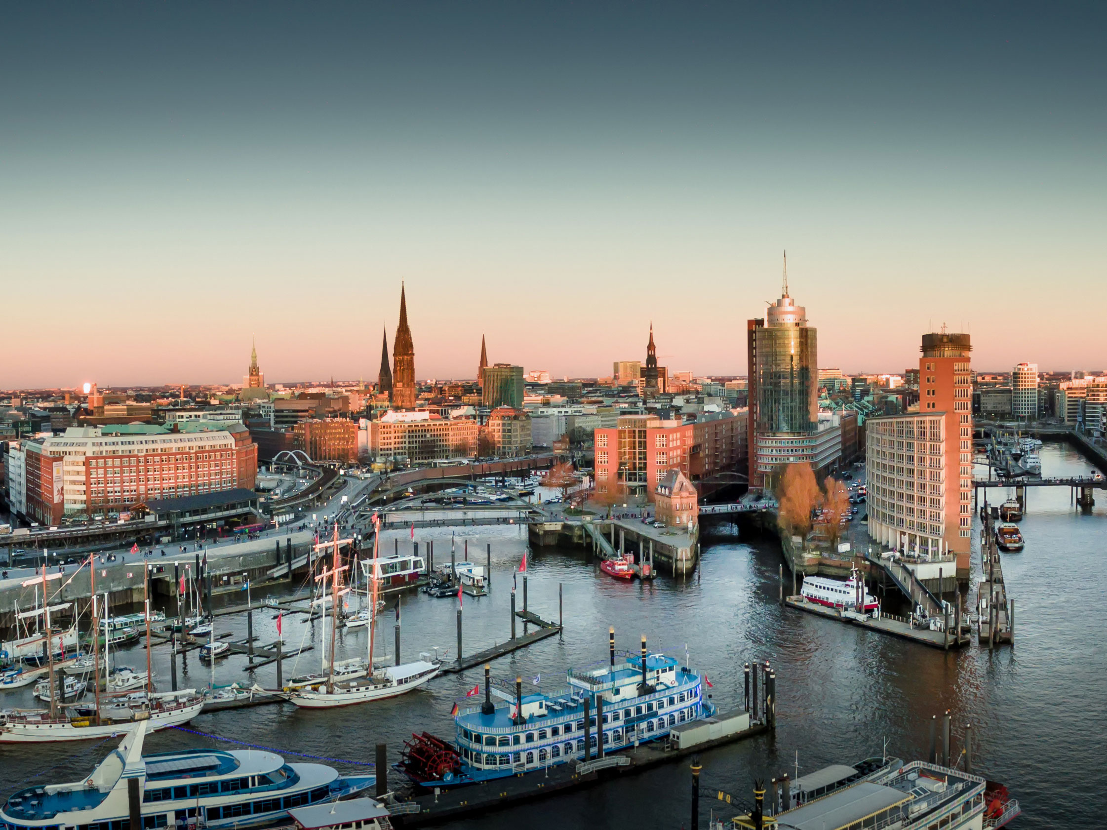 Panoramablick auf den Hamburger Hafen mit Segelschiffen, modernen Gebäuden und Kirchtürmen im warmen Licht der Abendsonne.